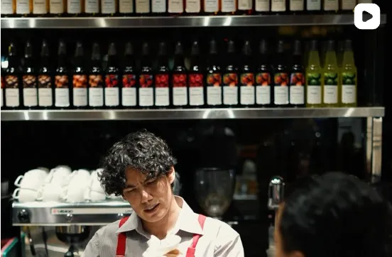 The Lokmaco baristasi qizil bretellarda, orqasida siroplar devori